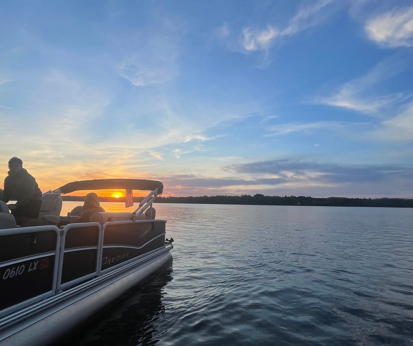 A pontoon is leaving the frame of the photo in the bottom left corner of the photo. You can see two people on the boat. A forested shoreline is in the distance and the sun is descending into the horizon behind the boat.